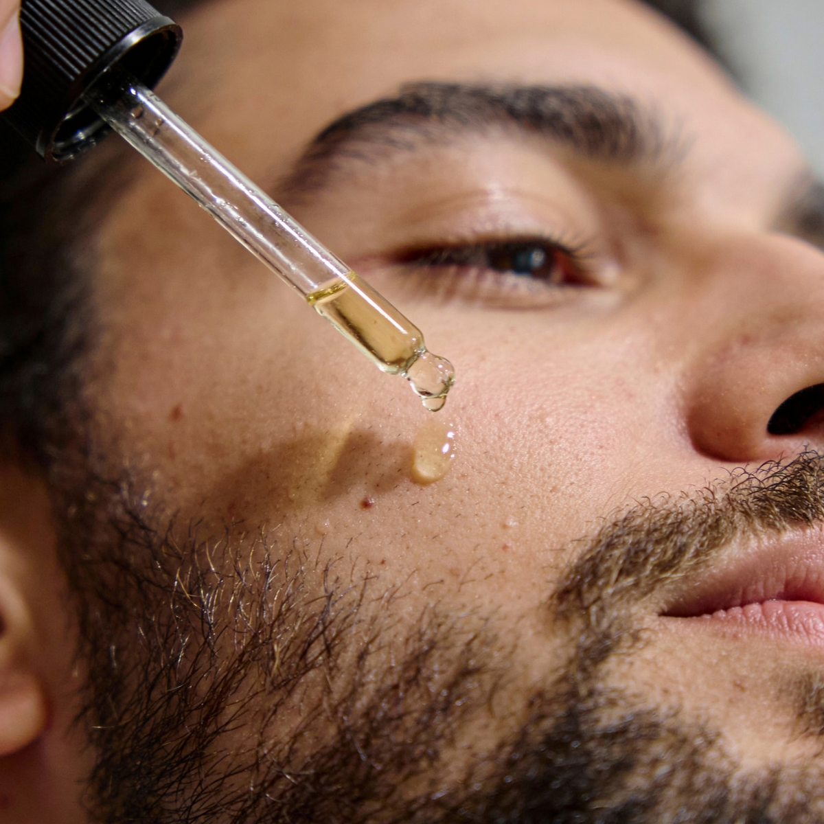 Close-up of a man applying a dropper of RAZOR MD Beard Oil to his face.